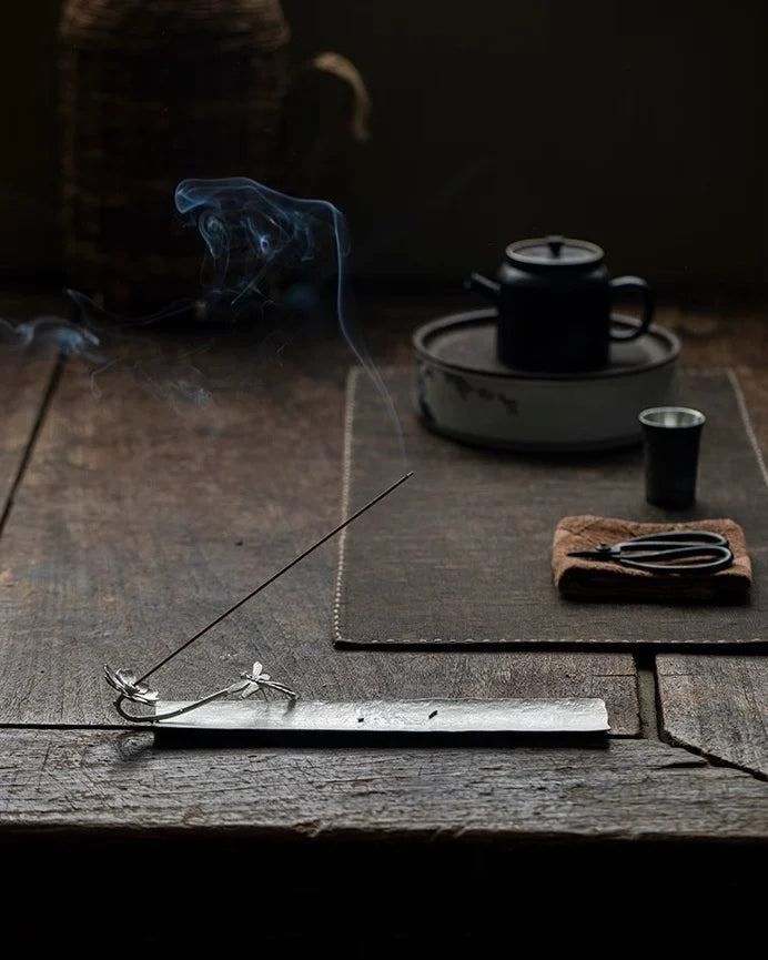 A handcrafted metal lotus incense holder with tea-making setup with teapot, cups, and Feng Shui incense on a wooden surface.