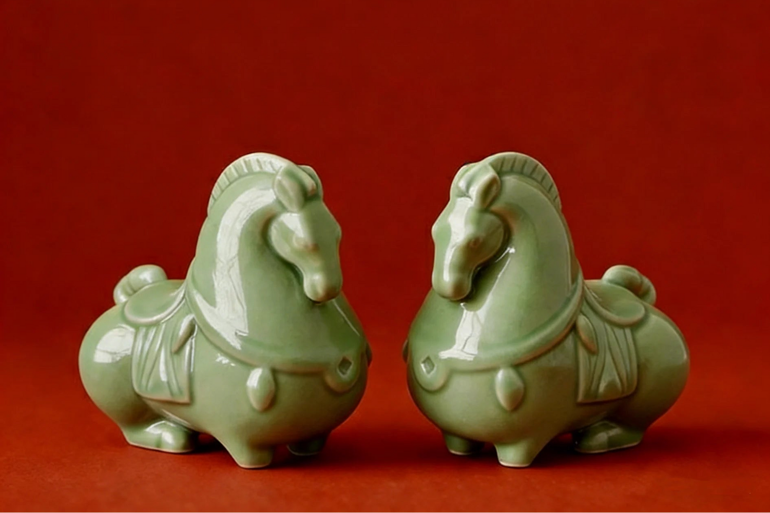 Two green ceramic horse figurines on a red background with an embroidered cloth at the top.
