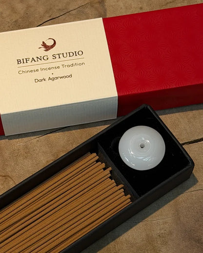 Bifang Studio Dark Agarwood Chinese Incense set, featuring a black tray with incense sticks and a white jade-style incense holder, shown next to its red and cream packaging box on a stone surface.