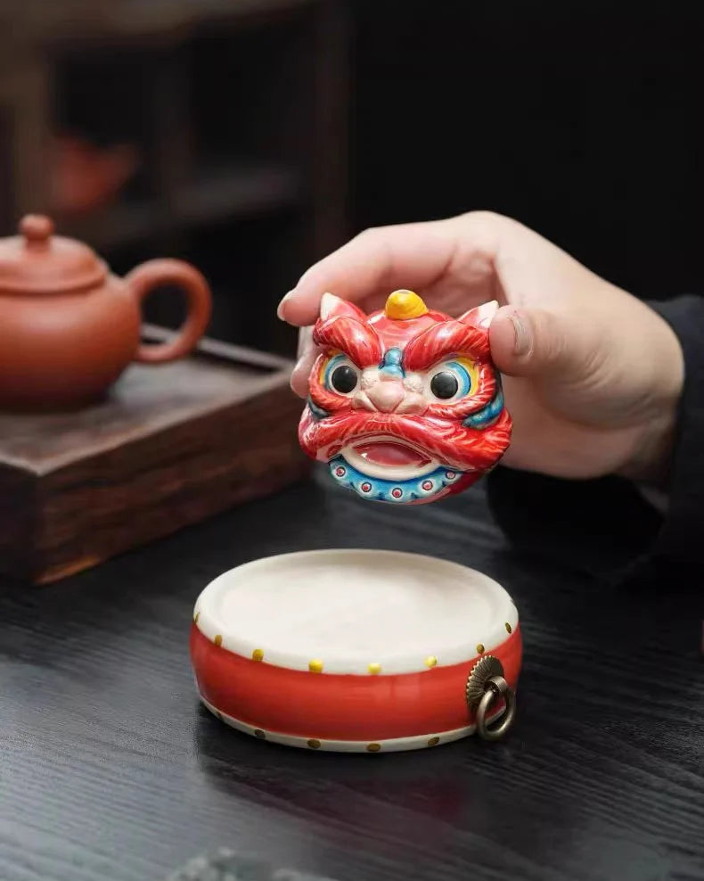 Hand holding a red ceramic lion-shaped incense holder with a teapot in the background