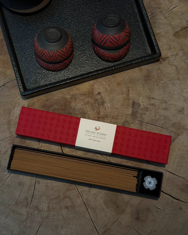 A boxed incense set displayed on a wood surface, including a red patterned incense box, a tray of brown incense sticks, and a small white flower-shaped incense holder. A second red box is placed behind the first on a black tray.