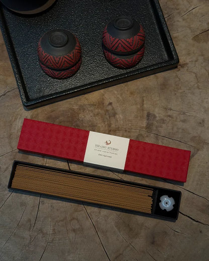 A boxed incense set displayed on a wood surface, including a red patterned incense box, a tray of brown incense sticks, and a small white flower-shaped incense holder. A second red box is placed behind the first on a black tray.