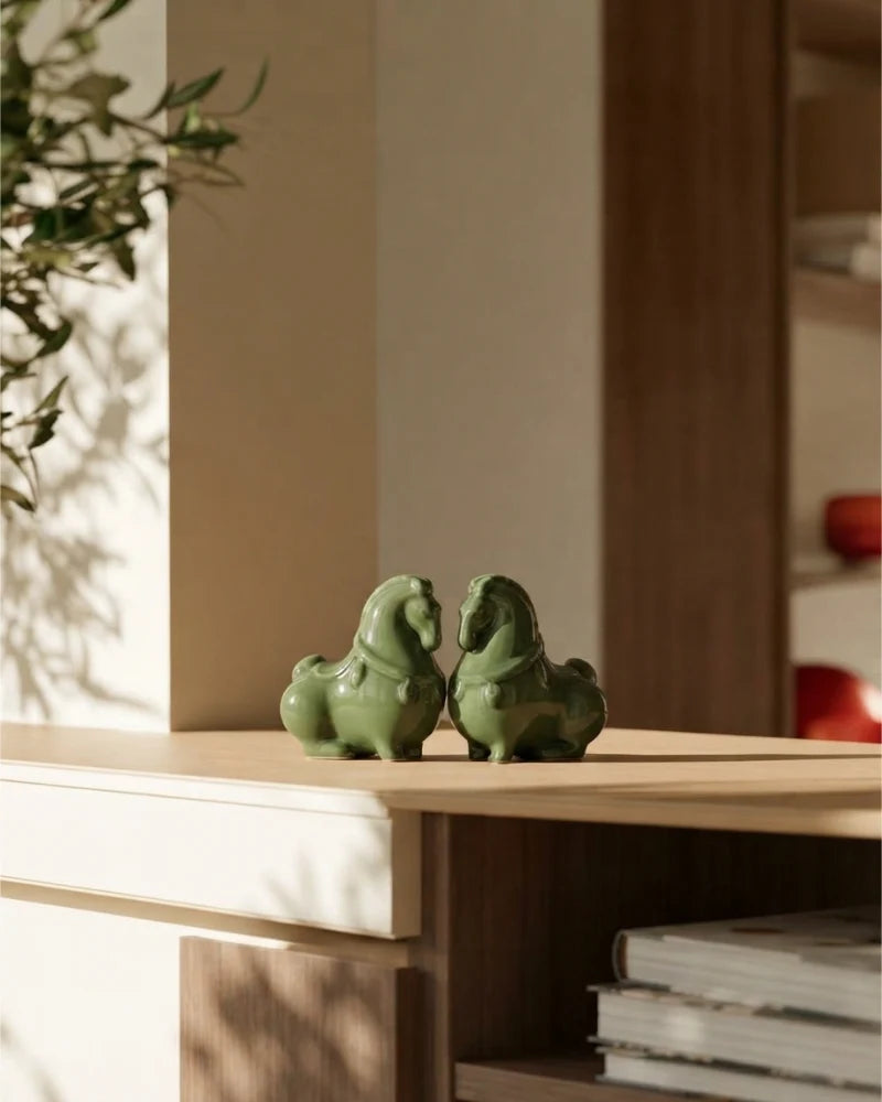 Two small glossy green horse figurines placed on a light wood shelf in a warm, sunlit interior setting.
