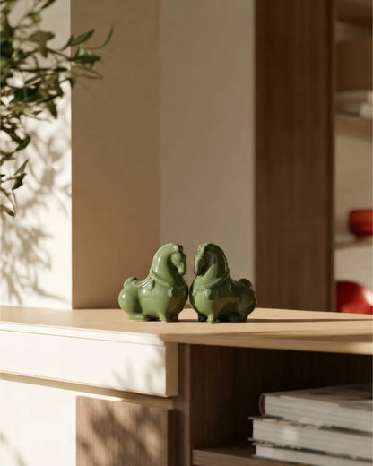 Two small glossy green horse figurines placed on a light wood shelf in a warm, sunlit interior setting.