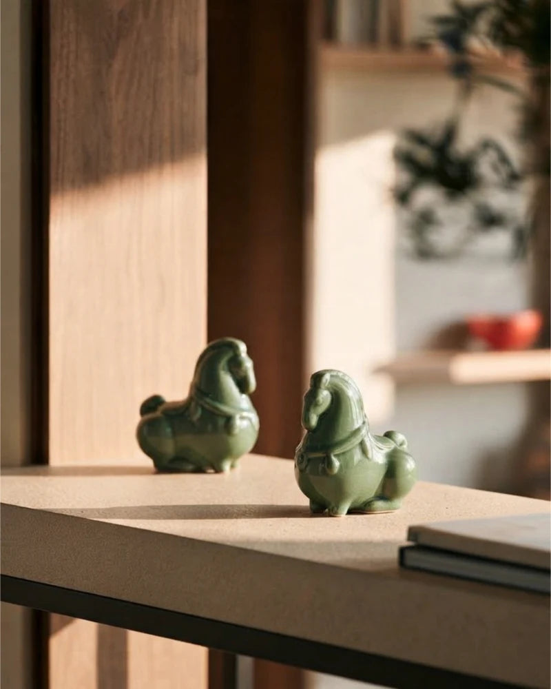 Two green celadon horse figurines displayed together on a sunlit wooden surface