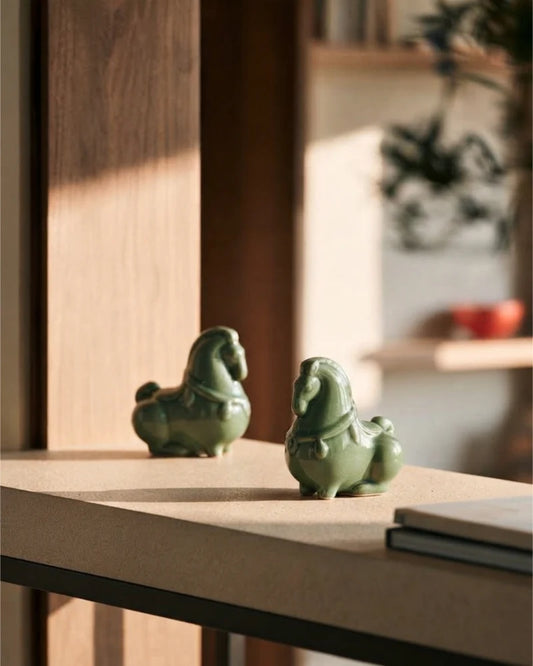 Two green celadon horse figurines displayed together on a sunlit wooden surface